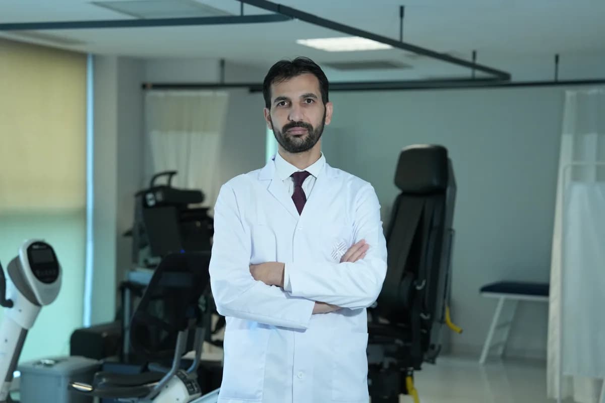 Beyaz önlüklü sakallı bir doktor, fizik tedavi cihazlarıyla dolu bir odada kollarını kavuşturmuş poz veriyor
