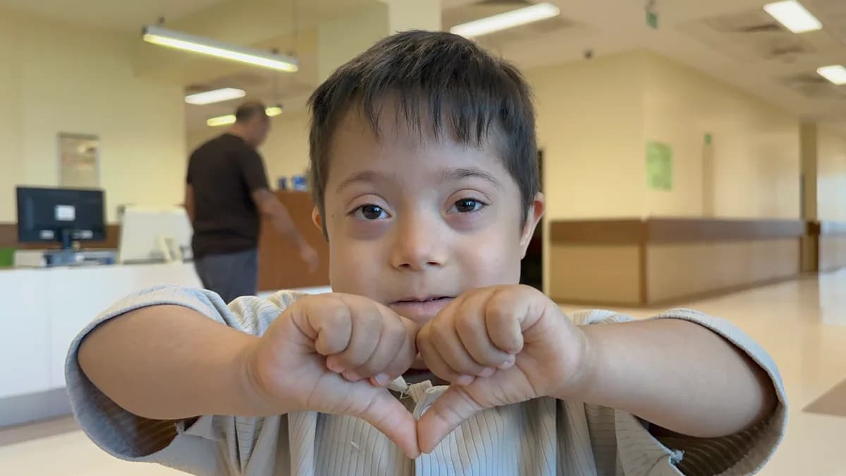 Down sendromlu bir çocuk elleriyle kalp işareti yaparak kameraya bakıyor